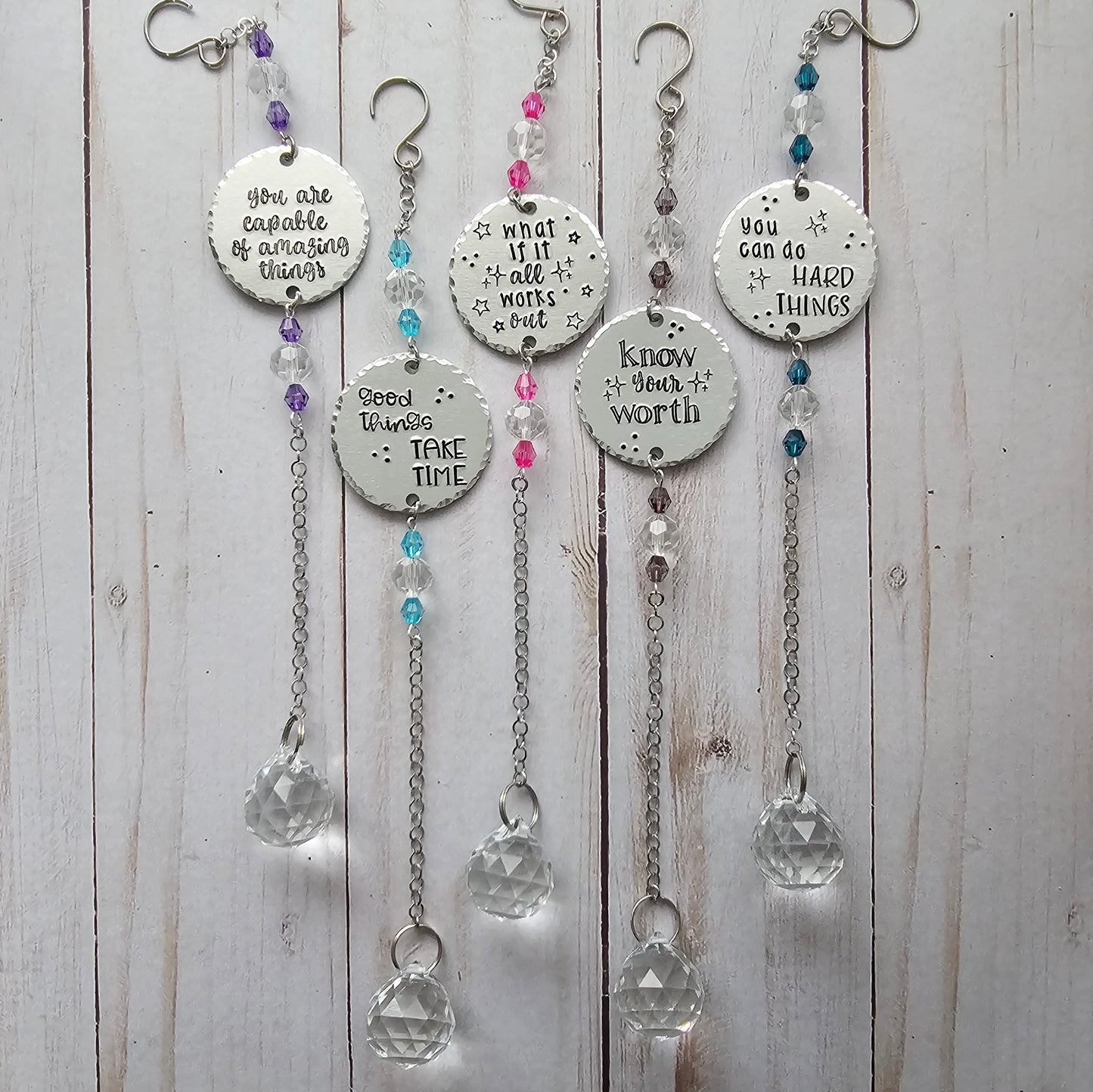 An image showing 5 suncatchers. Each suncatcher contains 2 sets of crystal beads, a round metal disc that is handstamped with a quote, chain, and a large crystal.