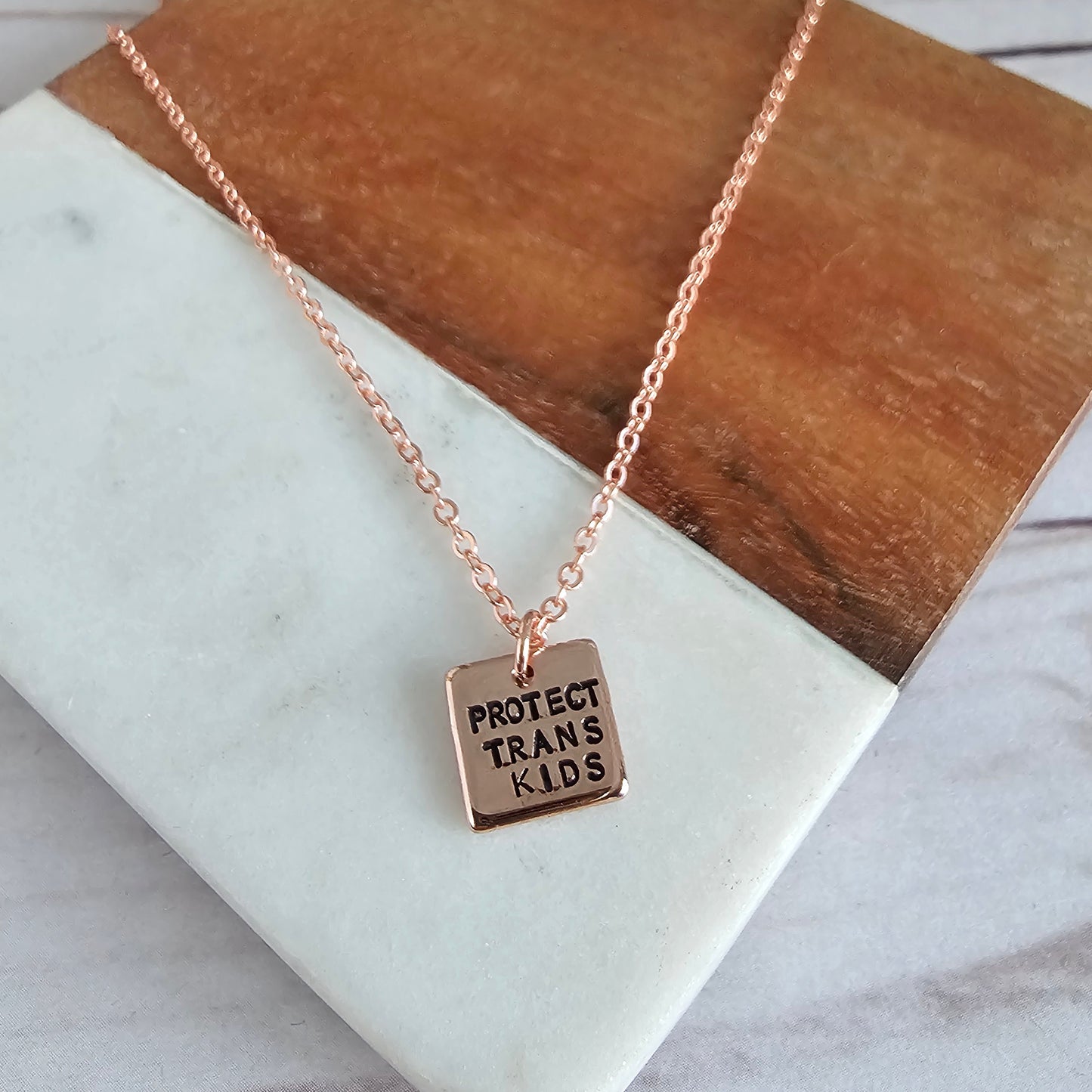 Rose Gold Square Charm that reads PROTECT TRANS KIDS, the charm is attached to a rose gold cable chain