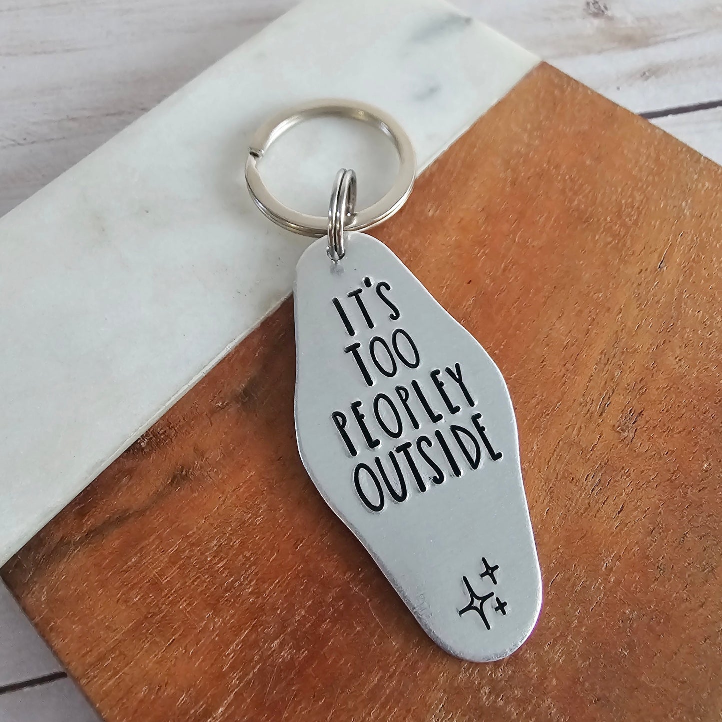 Silver keychain with 'It's Too Peopley Outside' text on a wooden surface