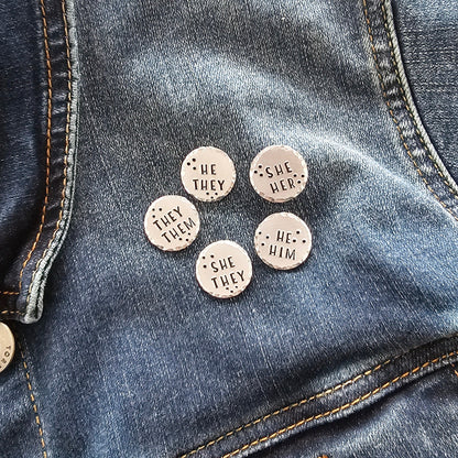 5 silver disc pins on a jean jacket. Each pin has different pronouns hand stamped onto the disc. They them, He They, She Her, She They, and He Him