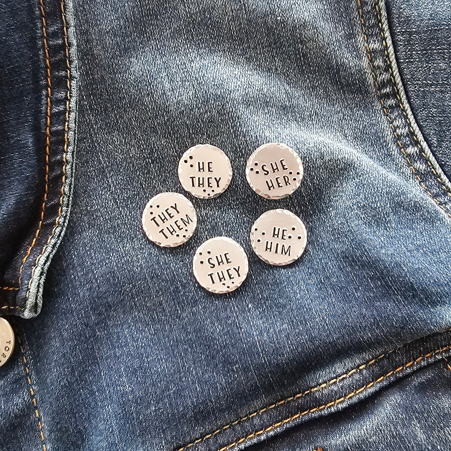5 silver disc pins on a jean jacket. Each pin has different pronouns hand stamped onto the disc. They them, He They, She Her, She They, and He Him