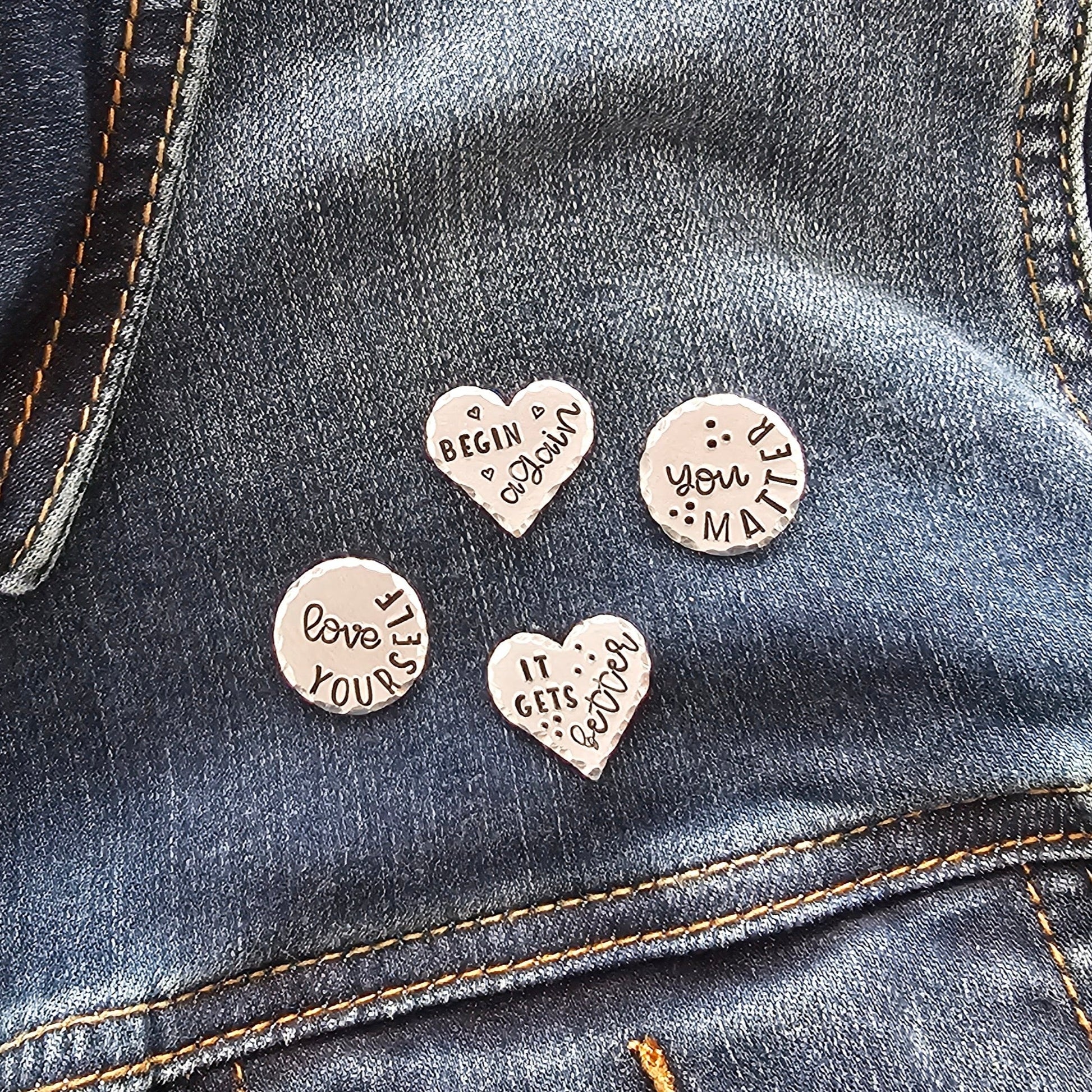 Jean jacket with 4 silver pins. 2 pins are round and have the messages love yourself and you matter. The heart pins read begin again and it gets better. The messages are handstamped onto the tags to leave a deep impression.