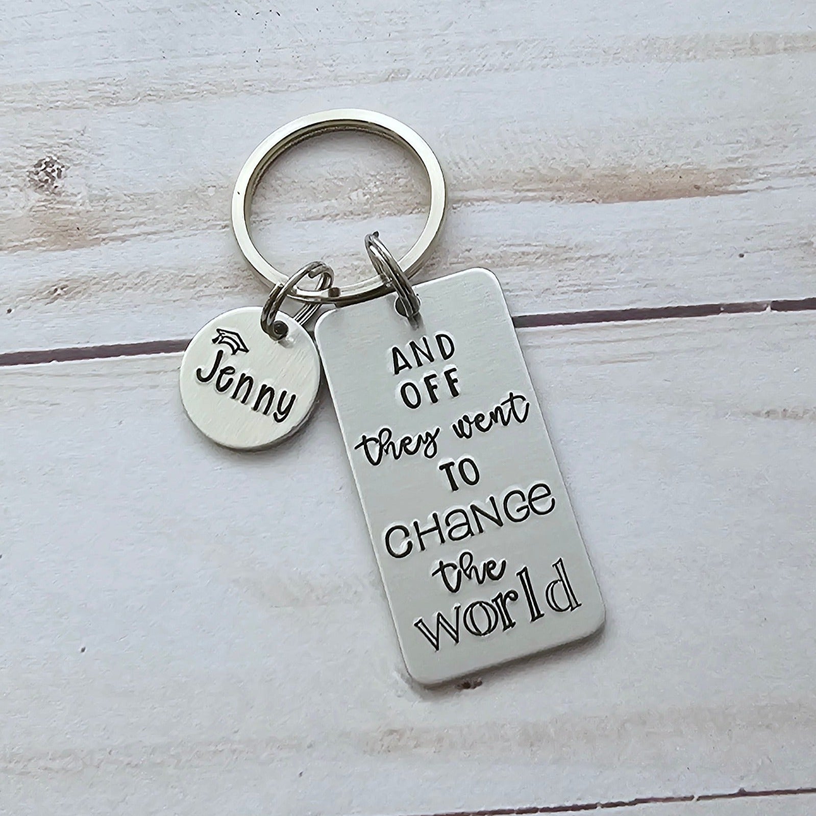 Silver rectangle keychain that reads and off they went to change the world. The keychain also has a round disc that you can customize the name, and the name has a graduation cap above it.