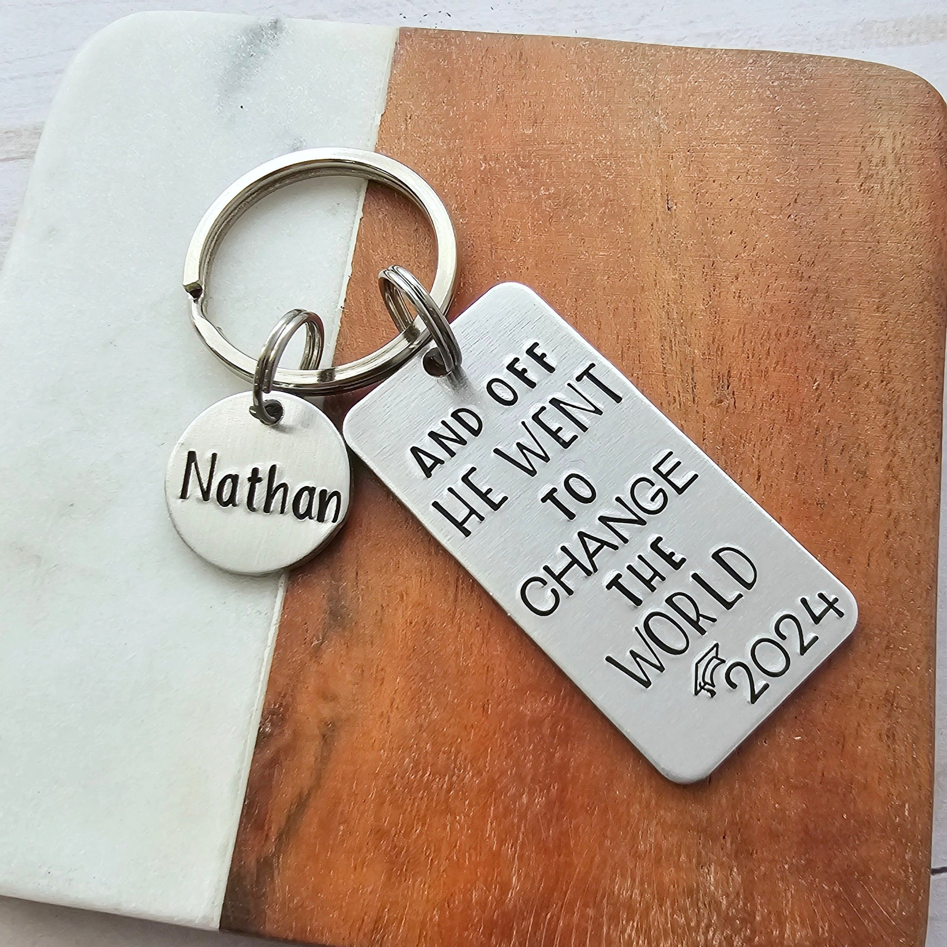Silver handstamped keychain that reads And off he went to change the world with the year 2024 and a grad cap. It also shows an optional round add on charm with a name of your choice. The hand stamped tag is a large rectangle.