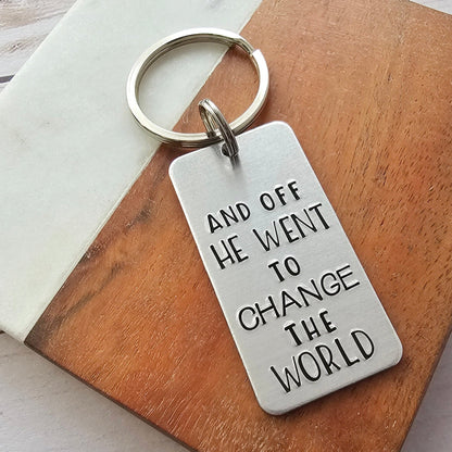 Silver rectangle tag that is hand stamped to read And off he went to change the world - the tag is attached to a split ring and key ring
