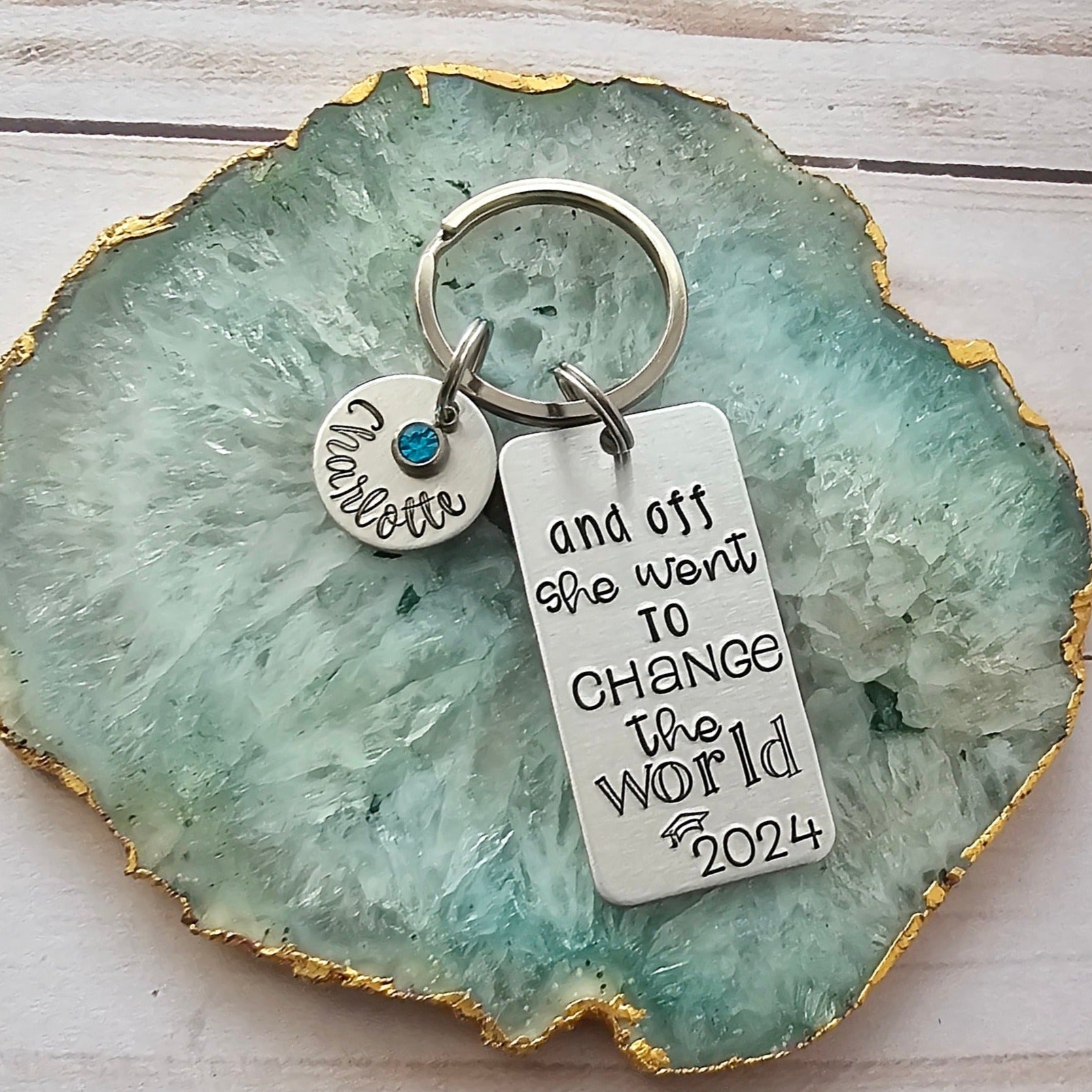 Silver rectangle keychain tag that reads And off she went to change the world with the year and a graduation cap. Also shown is an optional round disc with your choice of name and birthstone. Both charms are attached to a key ring.