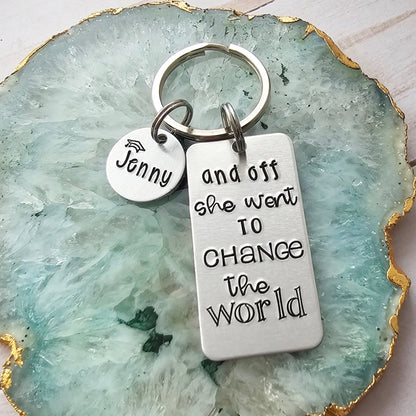 Silver rectangle keychain tag that is hand stamped to read and off she went to change the world. It also shows an optional round disc with a name and graduation cap. The tags are attached to a keyring.