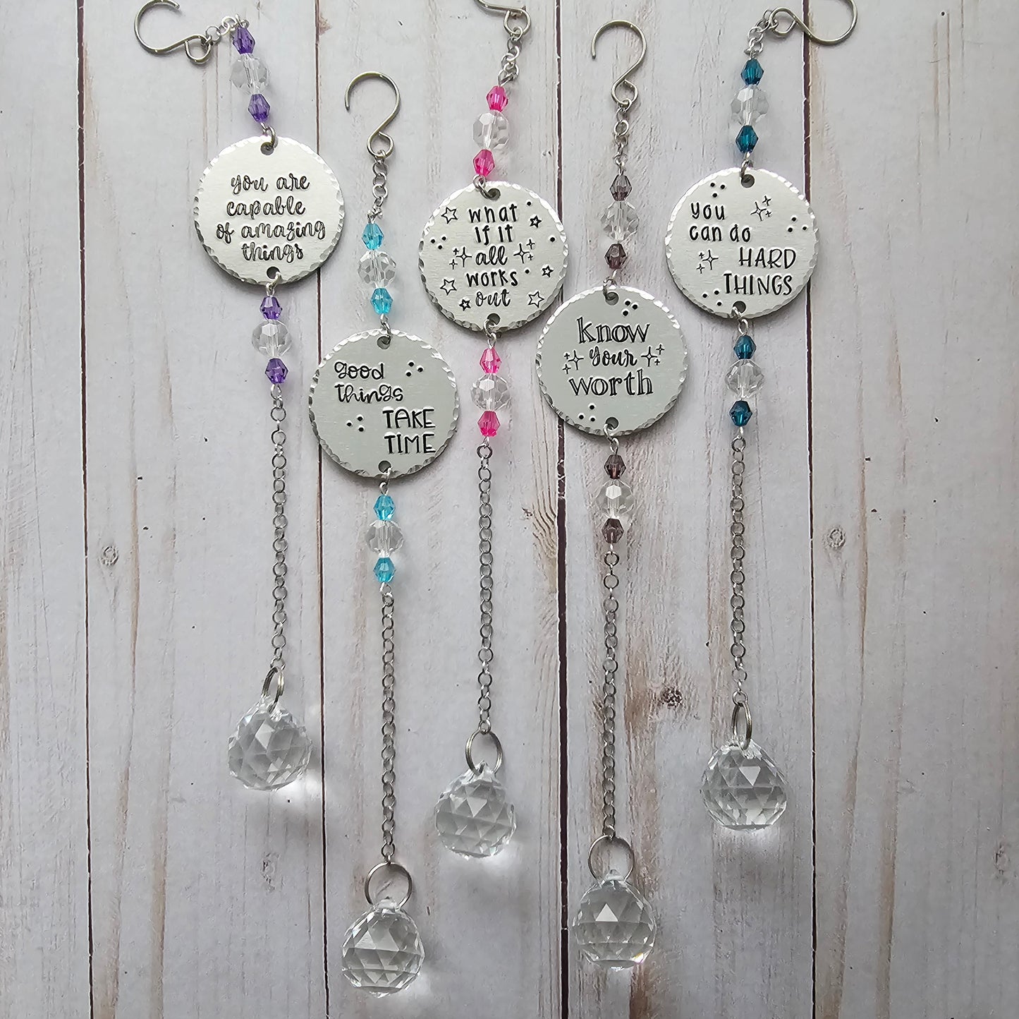 An image showing 5 suncatchers. Each suncatcher contains 2 sets of crystal beads, a round metal disc that is  handstamped with a quote, chain, and a large crystal.