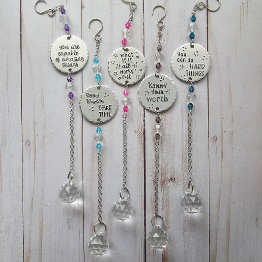 An image showing 5 suncatchers. Each suncatcher contains 2 sets of crystal beads, a round metal disc that is  handstamped with a quote, chain, and a large crystal.