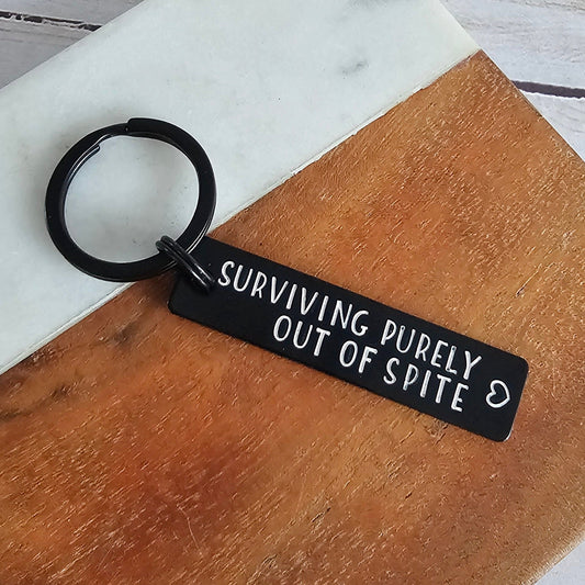 Keychain with 'Surviving Purely Out of Spite' text on a wooden block