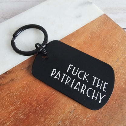 Black keychain with 'FUCK THE PATRIARCHY' text on a wooden surface