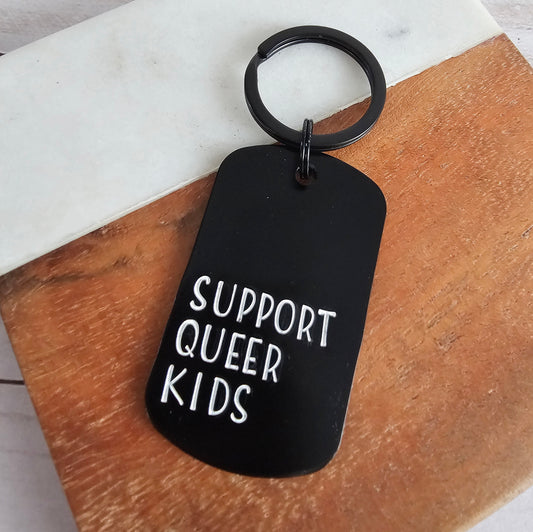 Black keychain with 'Support Queer Kids' text on a wooden surface