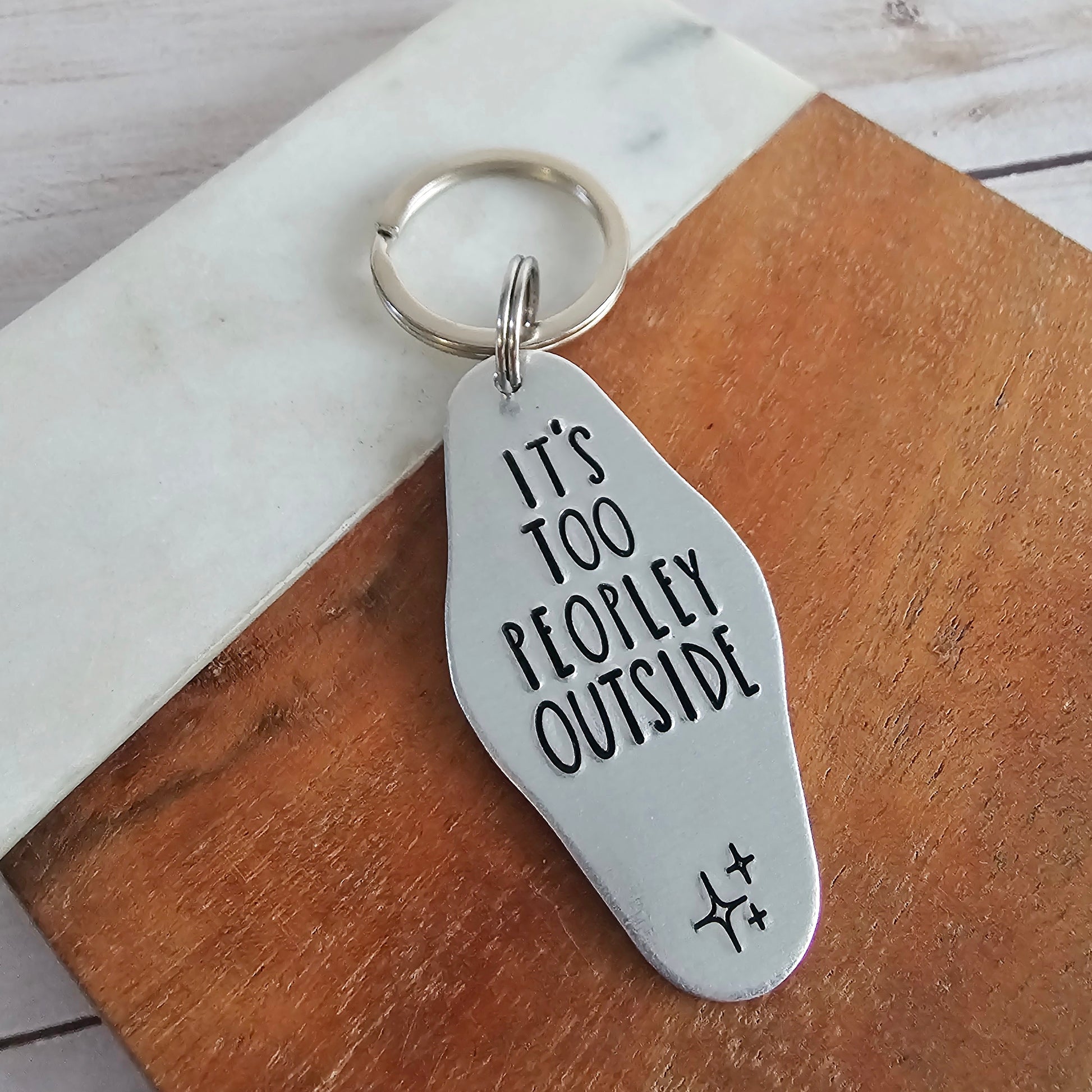 Silver keychain with 'It's Too Peopley Outside' text on a wooden surface