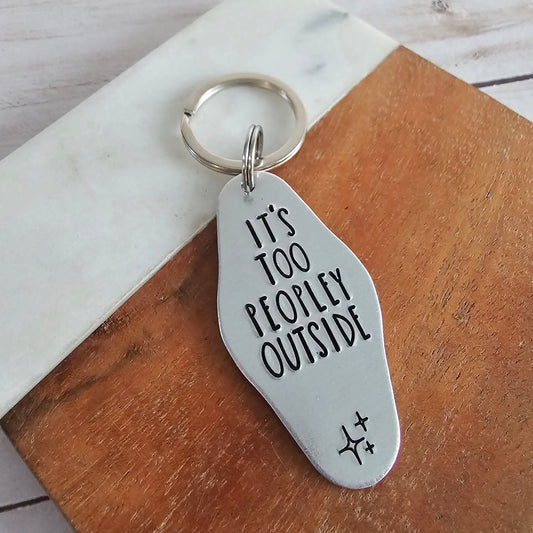 Silver keychain with 'It's Too Peopley Outside' text on a wooden surface