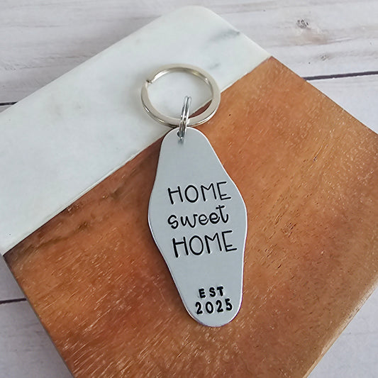 Silver keychain with 'HOME sweet HOME EST 2025' text on a wooden surface.