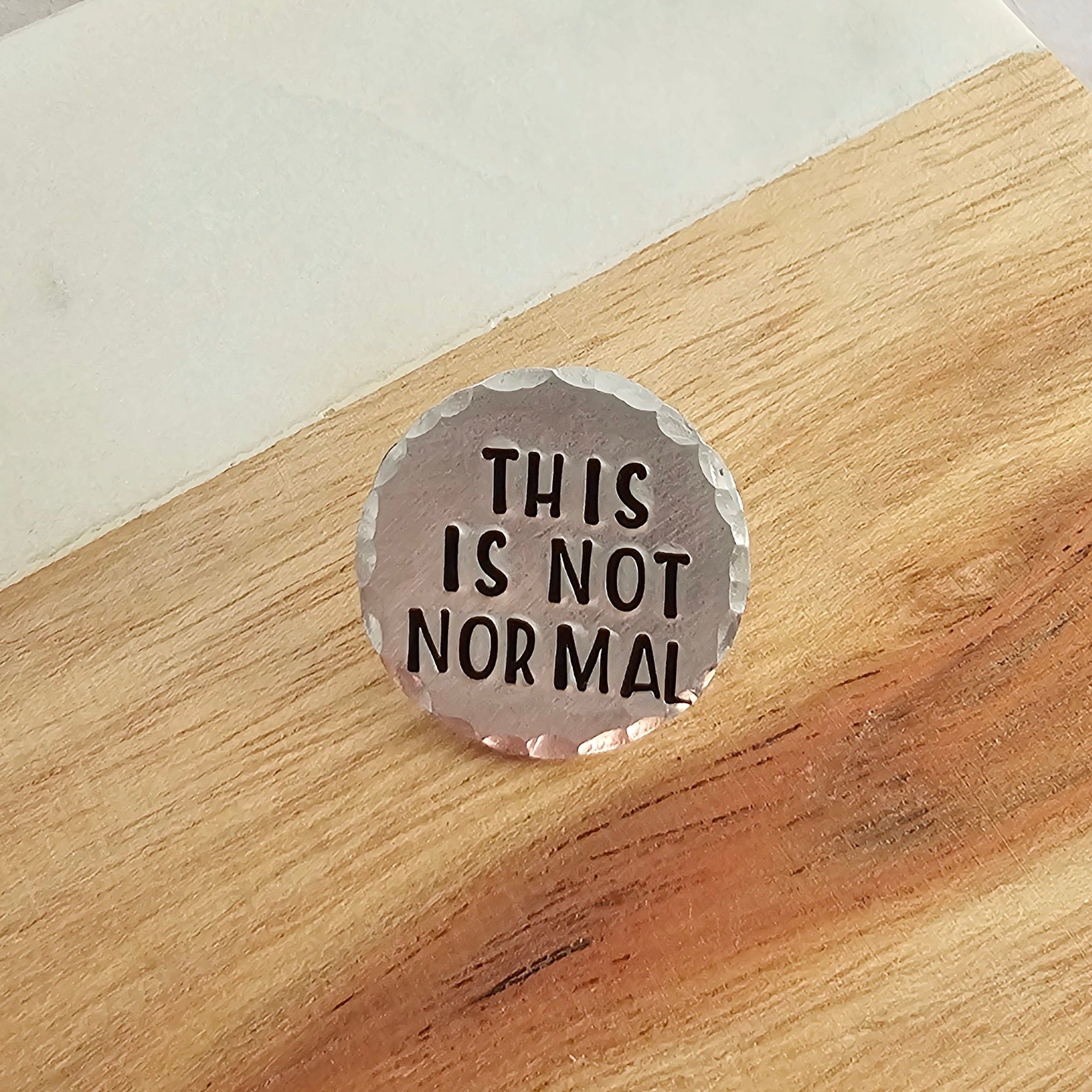 round silver disc with the text This Is not Normal with a wood and marble background