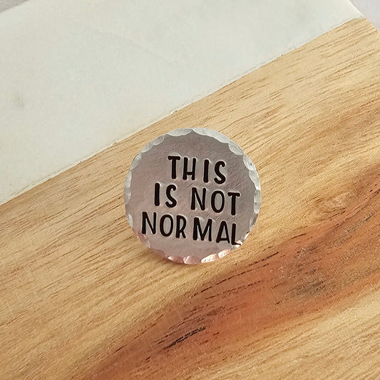 round silver disc with the text This Is not Normal with a wood and marble background
