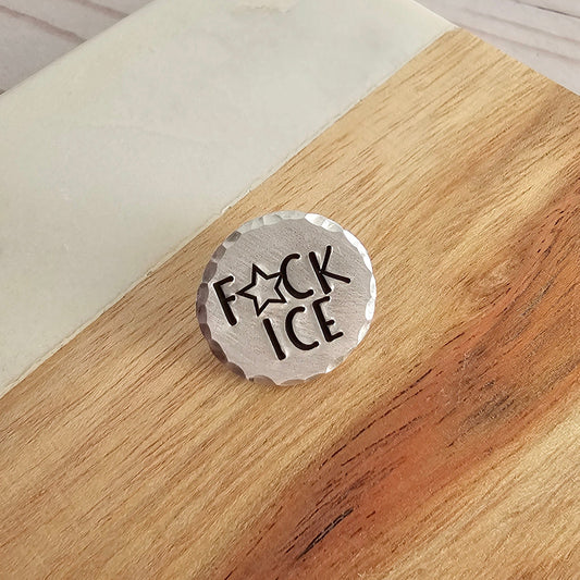 A silver round disc that is hand stamped to read F(star)CK ICE with a hammered edge. The Silver disc is placed on a wood and marble background.