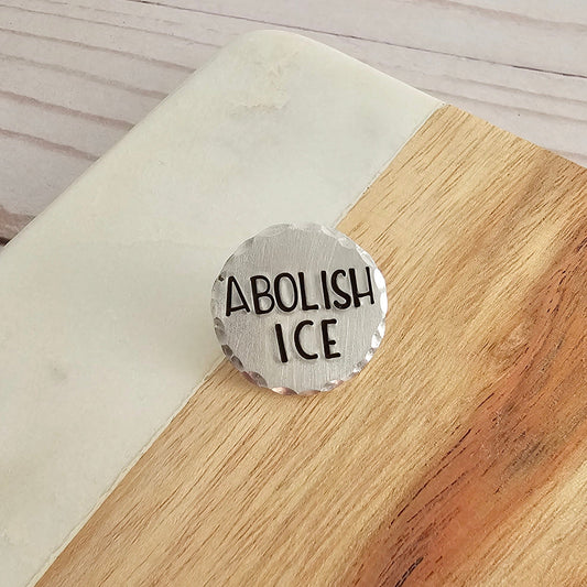 A round silver disc that is hand stamped to read ABOLISH ICE. The impression is filled in with black ink. The disc pin is placed on a wood and marble background.