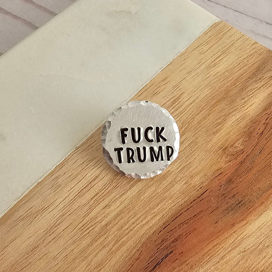 Round silver disc pin that reads FUCK TRUMP. 