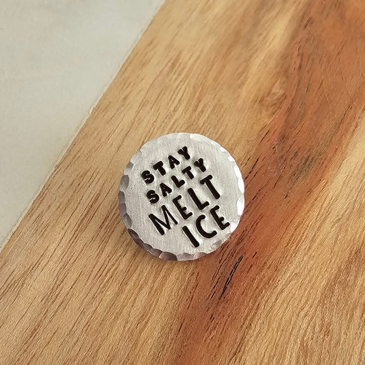 Round silver disc pin that reads stay salty melt ice and is displayed on a wood background