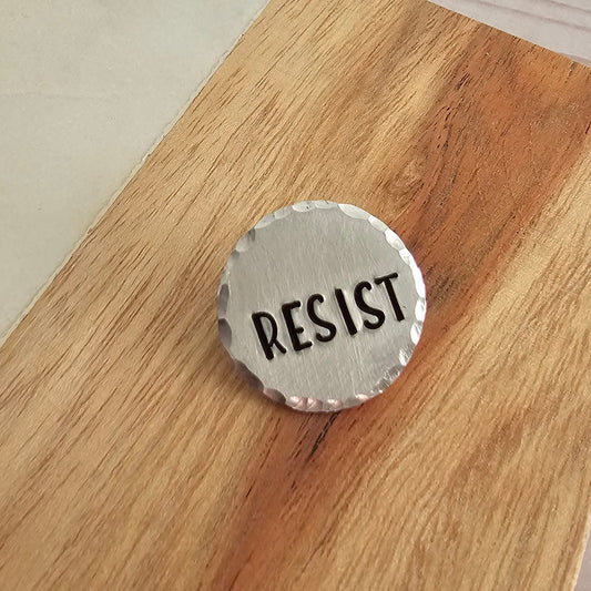 Round silver pin that reads RESIST and is displayed on a woodgrain background