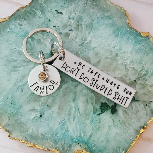 Silver rectangle tag that is hand stamped to read be safe, have fun, don't do stupid shit. It also shows a round charm with the name Taylor and a November birthstone. Both tags are attached to a key ring. They are placed on a teal colored tray with gold edge.