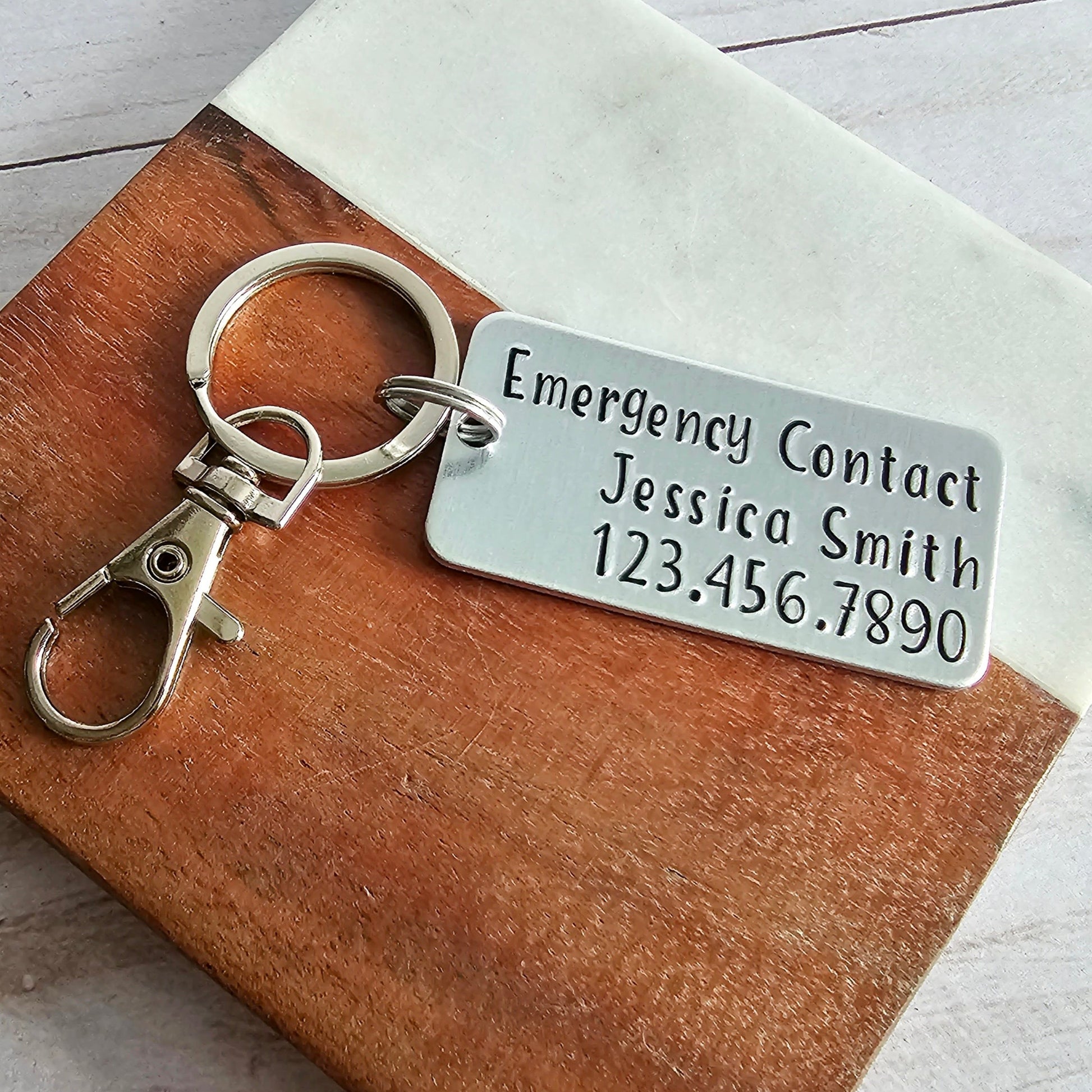 Silver metal keychain that is hand stamped to read Emergency Contact (first and last name) and (phone number)