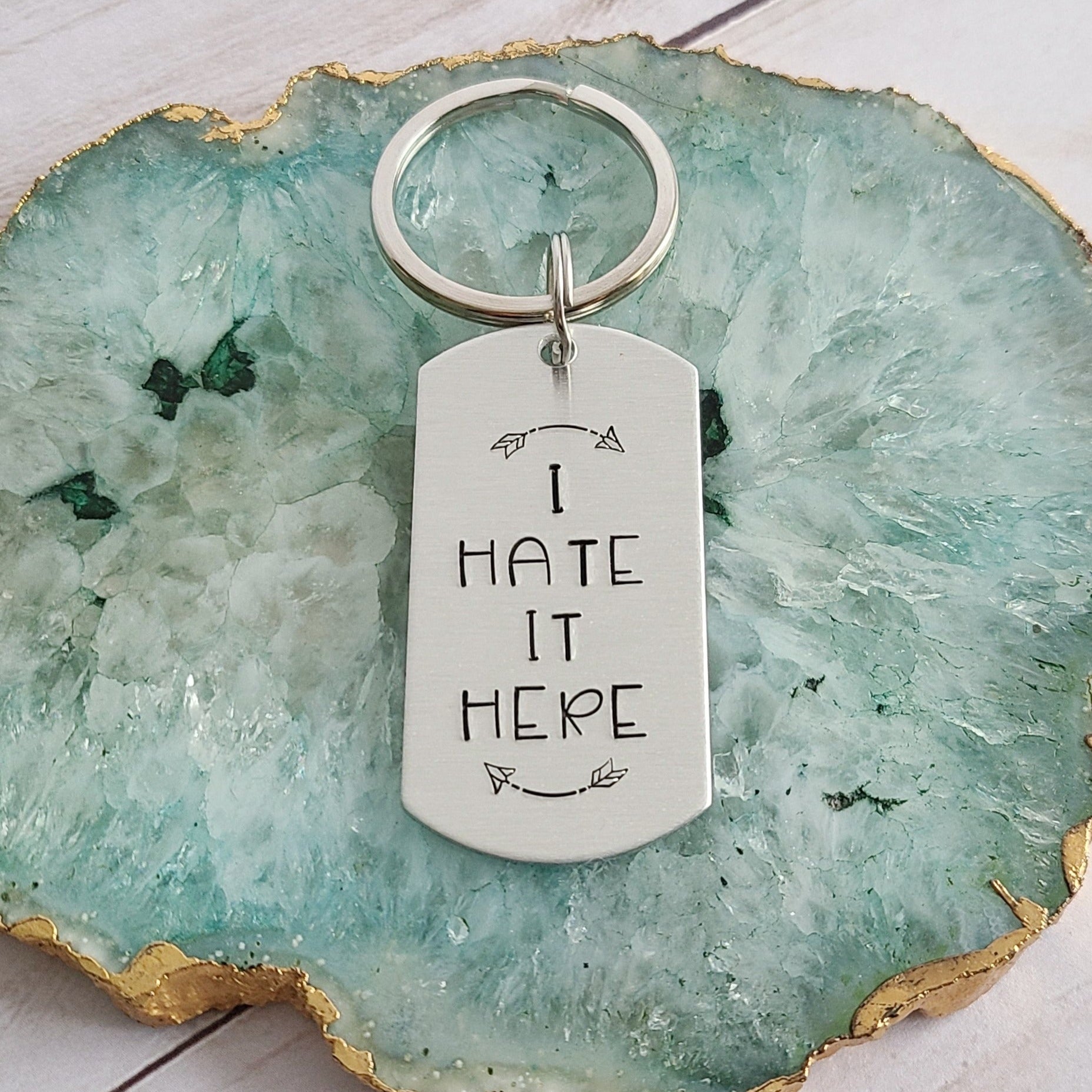 Silver colored dog taged shape tag that is hand stamped to read I Hate It Here with rounded arrows above and below the text. The Tag is attached to a key ring. The keychain is against a teal colored background.