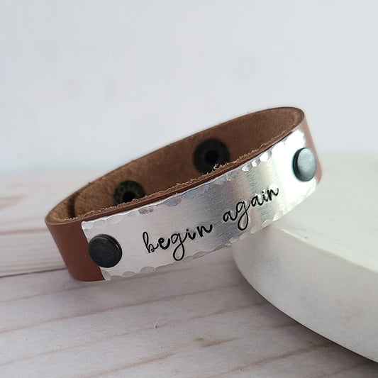 Natural Brown Leather Cuff Bracelet with a metal plate that is hand stamped to read begin again