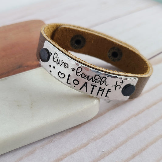 Leather cuff bracelet with a metal plate that is hand stamped to read live laugh loathe