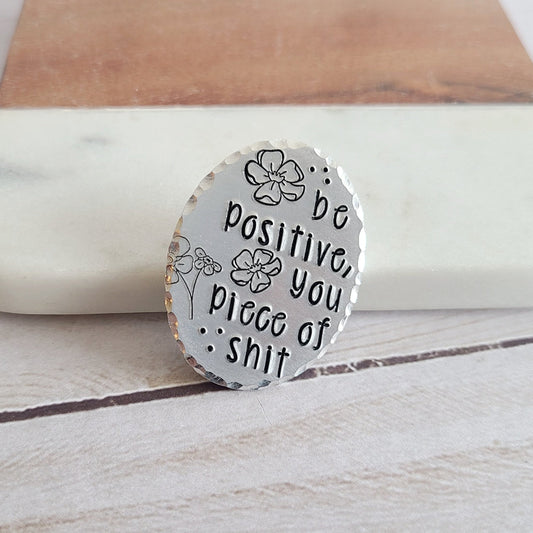 Silver oval pin that is hand stamped to read 'be positive, you piece of shit' with cute floral details
